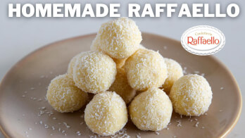 A pyramid of homemade Raffaello truffles sits on a beige plate. The truffles are round, covered in grated coconut, and presented on a light background. The text "HOMEMADE RAFFAELLO" is at the top, with a logo of Raffaello Confections on the right side.