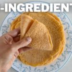 A hand is holding a folded piece of a flat, round food item on a blue and white floral plate. The text "2-INGREDIENT" is prominently displayed at the top of the image.