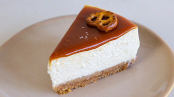 A slice of cheesecake topped with a caramel layer and garnished with a pretzel sits on a beige plate. The cheesecake has a crumbly graham cracker crust at the bottom.