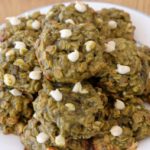 A plate of homemade cookies with a greenish hue, likely due to the inclusion of a unique ingredient like matcha or spinach. The cookies are studded with white chocolate chips and are served on a white plate with a blue rim, placed on a wooden surface.