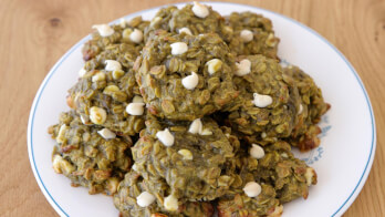 A plate of homemade cookies with a greenish hue, likely due to the inclusion of a unique ingredient like matcha or spinach. The cookies are studded with white chocolate chips and are served on a white plate with a blue rim, placed on a wooden surface.