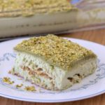 A slice of pistachio dessert, resembling a layered tiramisu, is served on a white plate with a blue floral pattern. The dessert has a greenish top layer with finely ground pistachios. In the background, the rest of the dessert is in a glass baking dish on a wooden table.