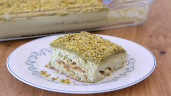 A slice of pistachio dessert, resembling a layered tiramisu, is served on a white plate with a blue floral pattern. The dessert has a greenish top layer with finely ground pistachios. In the background, the rest of the dessert is in a glass baking dish on a wooden table.