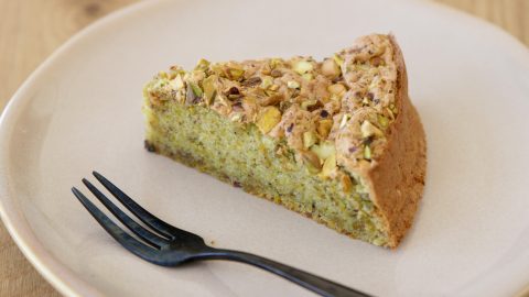 A slice of pistachio cake is served on a white plate with a black fork placed next to it. The cake has a light green hue and is topped with chopped pistachios. The background includes a light wooden surface.