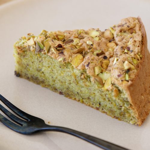 A slice of pistachio cake is served on a white plate with a black fork placed next to it. The cake has a light green hue and is topped with chopped pistachios. The background includes a light wooden surface.