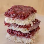 A stack of three raspberry coconut bars is centered on a light-colored plate. Each bar has a layer of raspberry jam on top of a shredded coconut base, showcasing their textured surfaces and vibrant red fruit topping. The background is out of focus.