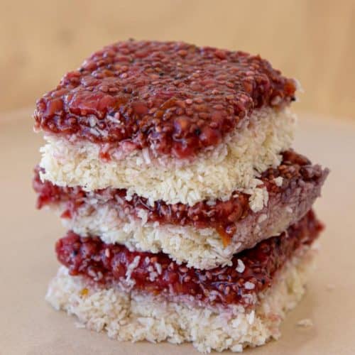 A stack of three raspberry coconut bars is centered on a light-colored plate. Each bar has a layer of raspberry jam on top of a shredded coconut base, showcasing their textured surfaces and vibrant red fruit topping. The background is out of focus.