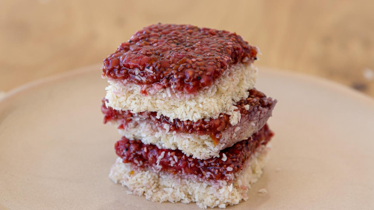 A stack of three raspberry coconut bars is centered on a light-colored plate. Each bar has a layer of raspberry jam on top of a shredded coconut base, showcasing their textured surfaces and vibrant red fruit topping. The background is out of focus.