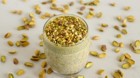 A glass jar filled with a greenish-yellow pistachio-flavored pudding topped with chopped pistachios, placed on a light surface scattered with whole and chopped pistachio nuts. The pudding appears rich and creamy with visible chia seeds mixed in.