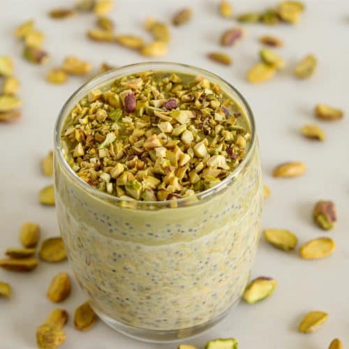 A glass jar filled with a greenish-yellow pistachio-flavored pudding topped with chopped pistachios, placed on a light surface scattered with whole and chopped pistachio nuts. The pudding appears rich and creamy with visible chia seeds mixed in.