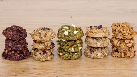 Five stacks of homemade cookies sit on a wooden surface, each stack featuring a different variety: dark chocolate, oatmeal raisin, matcha white chocolate, chocolate chip, and oatmeal peanut butter. The cookies have a rustic, hand-formed appearance.