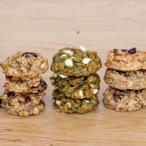 Five stacks of homemade cookies sit on a wooden surface, each stack featuring a different variety: dark chocolate, oatmeal raisin, matcha white chocolate, chocolate chip, and oatmeal peanut butter. The cookies have a rustic, hand-formed appearance.