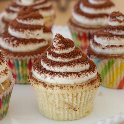 A plate of cupcakes is topped with swirled white frosting and dusted with cocoa powder. The cupcakes are in colorful, striped paper liners and are arranged closely together on a white plate. The background is out of focus.