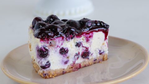 A slice of blueberry cheesecake on a beige plate. The cheesecake has a graham cracker crust, creamy filling with blueberries mixed in, and a glossy blueberry topping. The background is out of focus, featuring a hint of a white pedestal cake stand.