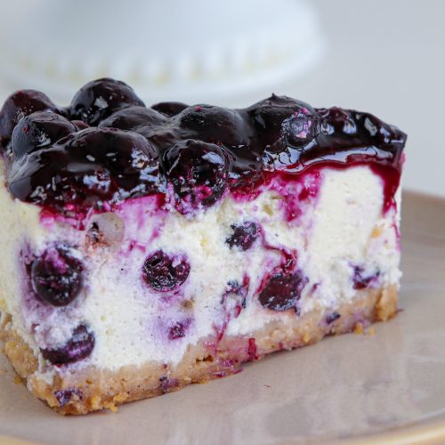 A slice of blueberry cheesecake on a beige plate. The cheesecake has a graham cracker crust, creamy filling with blueberries mixed in, and a glossy blueberry topping. The background is out of focus, featuring a hint of a white pedestal cake stand.