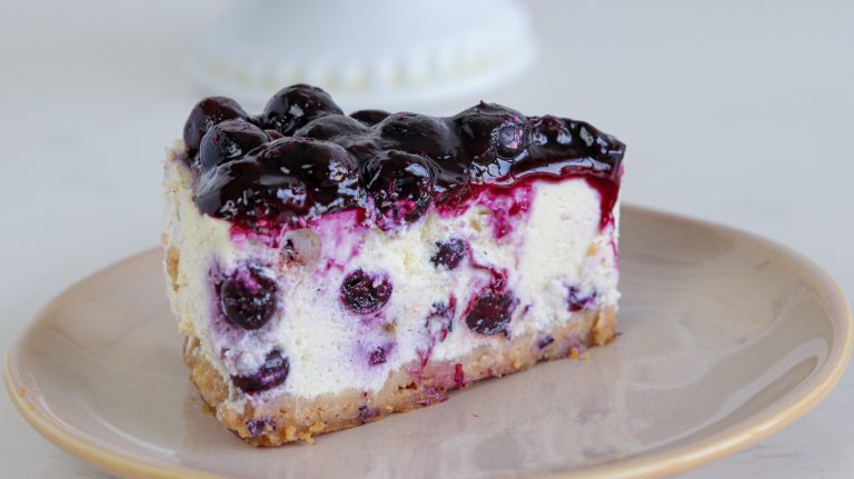 A slice of blueberry cheesecake on a beige plate. The cheesecake has a graham cracker crust, creamy filling with blueberries mixed in, and a glossy blueberry topping. The background is out of focus, featuring a hint of a white pedestal cake stand.