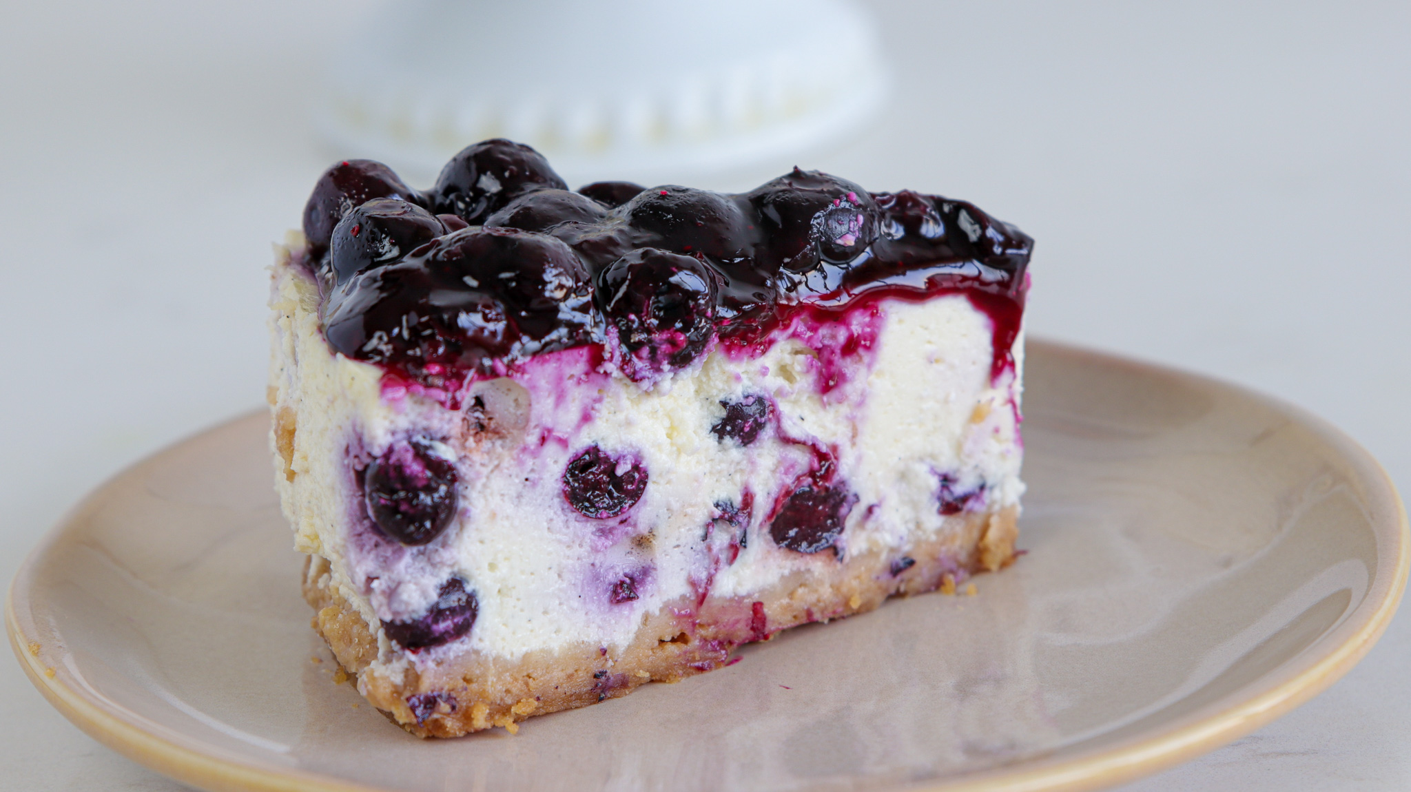 A slice of blueberry cheesecake on a beige plate. The cheesecake has a graham cracker crust, creamy filling with blueberries mixed in, and a glossy blueberry topping. The background is out of focus, featuring a hint of a white pedestal cake stand.