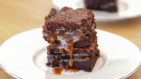 A close-up of a decadent chocolate brownie stack with rich caramel drizzle oozing between the layers, served on a white plate. Another brownie is slightly blurred in the background on another plate. The dessert sits on a light wooden surface.