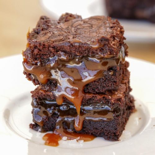 A close-up of a decadent chocolate brownie stack with rich caramel drizzle oozing between the layers, served on a white plate. Another brownie is slightly blurred in the background on another plate. The dessert sits on a light wooden surface.