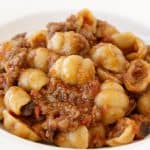 A plate of orecchiette pasta topped with a savory meat sauce. The pasta is covered with a rich, reddish-brown sauce that includes ground meat, giving a hearty and flavorful appearance. The dish is served in a white ceramic bowl with a decorative rim.