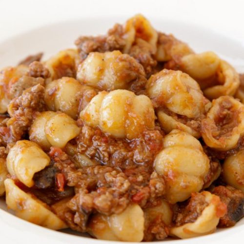 A plate of orecchiette pasta topped with a savory meat sauce. The pasta is covered with a rich, reddish-brown sauce that includes ground meat, giving a hearty and flavorful appearance. The dish is served in a white ceramic bowl with a decorative rim.