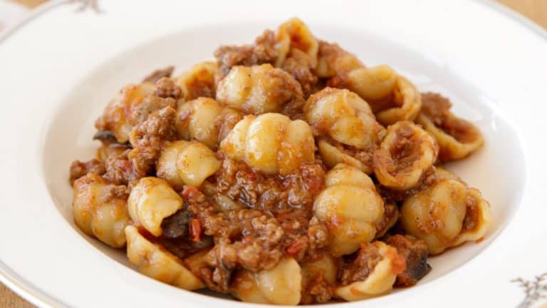 A plate of orecchiette pasta topped with a savory meat sauce. The pasta is covered with a rich, reddish-brown sauce that includes ground meat, giving a hearty and flavorful appearance. The dish is served in a white ceramic bowl with a decorative rim.