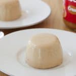 Two creamy panna cotta desserts are served on white plates placed on a wooden surface. Two silver spoons lie beside the plates. In the background, a jar of Biscoff cookie spread is partially visible, suggesting the desserts might be flavored with it.