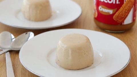 Two creamy panna cotta desserts are served on white plates placed on a wooden surface. Two silver spoons lie beside the plates. In the background, a jar of Biscoff cookie spread is partially visible, suggesting the desserts might be flavored with it.