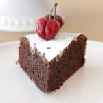 A triangular slice of chocolate cake is placed on a white plate. The cake is topped with a dusting of powdered sugar and two red cherries. The background includes a blurred white cake stand.