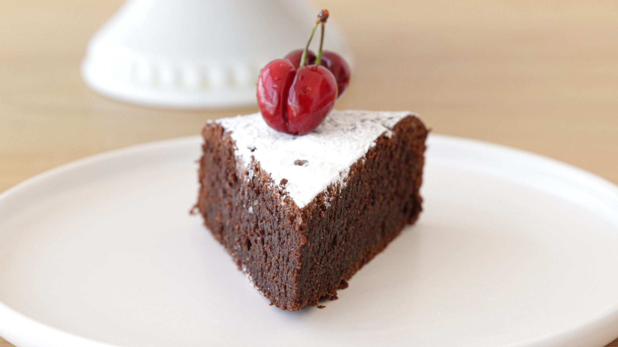 A triangular slice of chocolate cake is placed on a white plate. The cake is topped with a dusting of powdered sugar and two red cherries. The background includes a blurred white cake stand.