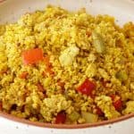 A ceramic bowl filled with a colorful mix of bulgur pilaf. The dish features golden-colored bulgur grains, red bell pepper pieces, and small chunks of vegetables, creating a vibrant and appealing presentation. The bowl rests on a wooden surface.