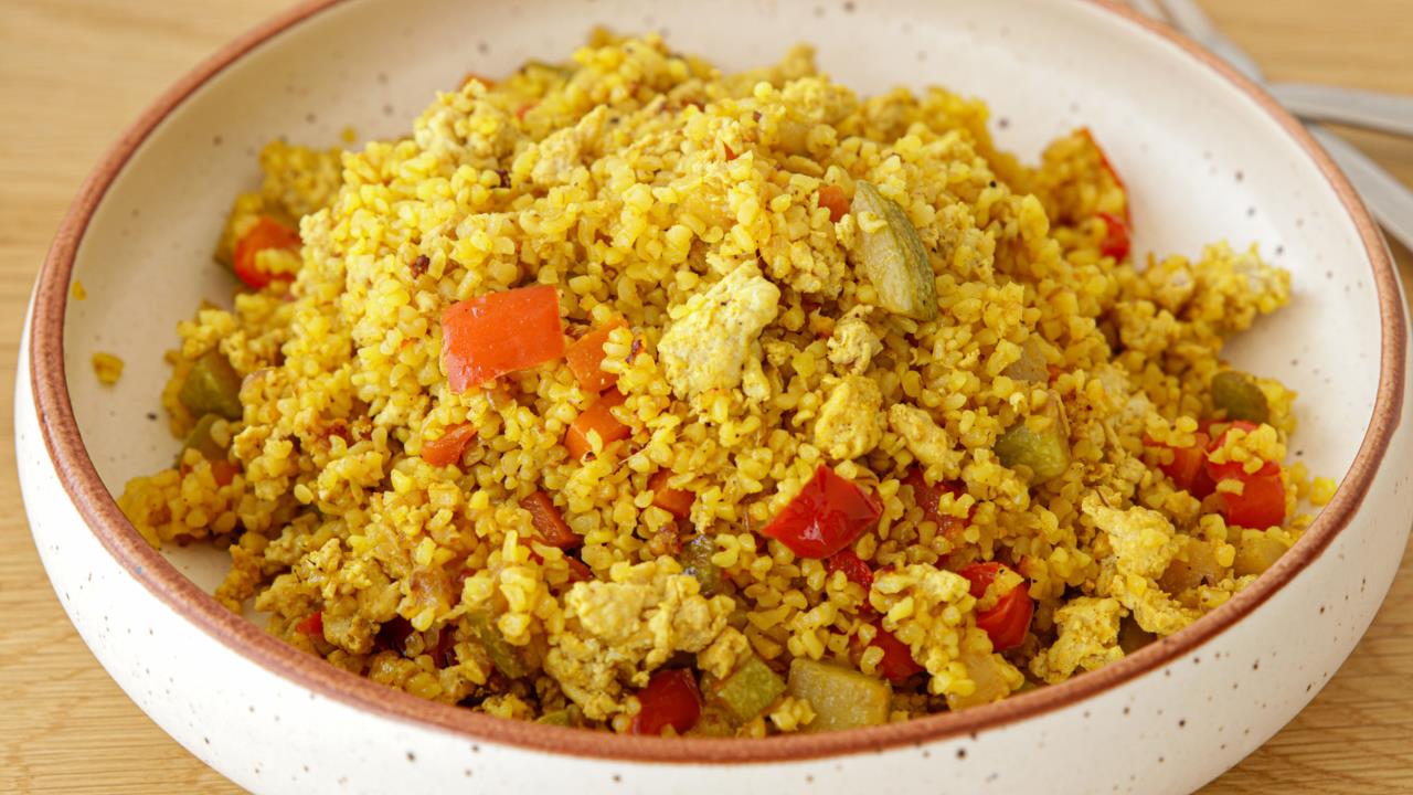 A ceramic bowl filled with a colorful mix of bulgur pilaf. The dish features golden-colored bulgur grains, red bell pepper pieces, and small chunks of vegetables, creating a vibrant and appealing presentation. The bowl rests on a wooden surface.