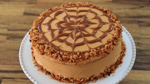 A round cake with a smooth caramel-colored frosting is decorated with a web-like swirl of caramel on top. The edge of the cake is garnished with crumbly brown bits, and it is placed on a white plate with a wooden surface in the background.