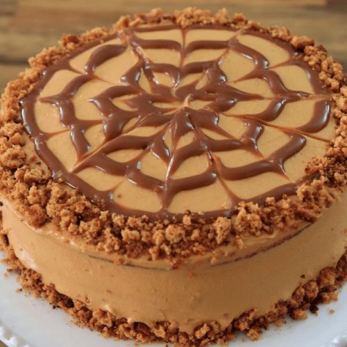 A round cake with a smooth caramel-colored frosting is decorated with a web-like swirl of caramel on top. The edge of the cake is garnished with crumbly brown bits, and it is placed on a white plate with a wooden surface in the background.