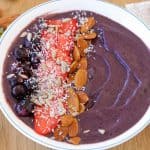 A vibrant acai bowl is topped with fresh strawberries, blueberries, almonds, sliced strawberries, and shredded coconut. It sits on a wooden table with a red and white cloth to the right and scattered strawberries and almonds around.