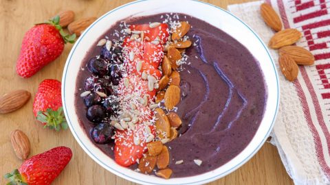 A vibrant acai bowl is topped with fresh strawberries, blueberries, almonds, sliced strawberries, and shredded coconut. It sits on a wooden table with a red and white cloth to the right and scattered strawberries and almonds around.