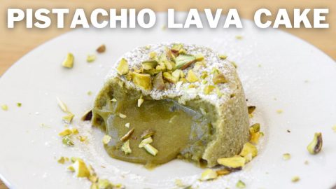 A pistachio lava cake sits on a wooden surface, with molten pistachio filling oozing out from the center. The cake is topped with chopped pistachios and a dusting of powdered sugar. The words "PISTACHIO LAVA CAKE" are displayed prominently above the dessert.