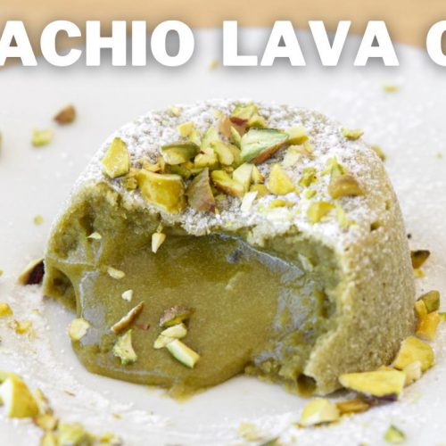 A pistachio lava cake sits on a wooden surface, with molten pistachio filling oozing out from the center. The cake is topped with chopped pistachios and a dusting of powdered sugar. The words "PISTACHIO LAVA CAKE" are displayed prominently above the dessert.