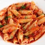 A white plate contains a serving of penne pasta topped with a rich tomato sauce and garnished with finely chopped parsley. The sauce is thick and chunky, covering the pasta evenly. The dish is placed on a light-colored surface.