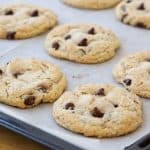 A baking tray lined with parchment paper holds freshly baked chocolate chip cookies. The cookies are golden brown with visible chocolate chips, and they are spread out evenly on the tray, suggesting they have just been taken out of the oven.