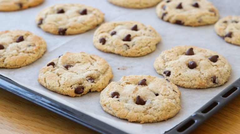 A baking tray lined with parchment paper holds freshly baked chocolate chip cookies. The cookies are golden brown with visible chocolate chips, and they are spread out evenly on the tray, suggesting they have just been taken out of the oven.