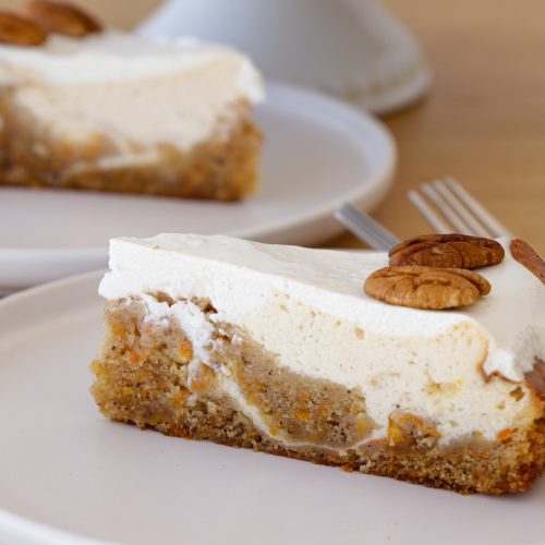 Two slices of carrot cake cheesecake are served on white plates. Each slice has a layer of carrot cake topped with a thick layer of cheesecake and is garnished with two pecan halves. A fork is placed next to each slice on the plates.