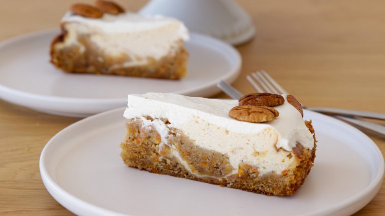 Two slices of carrot cake cheesecake are served on white plates. Each slice has a layer of carrot cake topped with a thick layer of cheesecake and is garnished with two pecan halves. A fork is placed next to each slice on the plates.