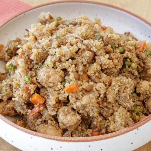 A bowl of quinoa fried rice with mixed vegetables like carrots, peas, and diced chicken. The dish is served in a speckled ceramic bowl placed on a wooden table, with a pink cloth napkin and silver utensils beside it.