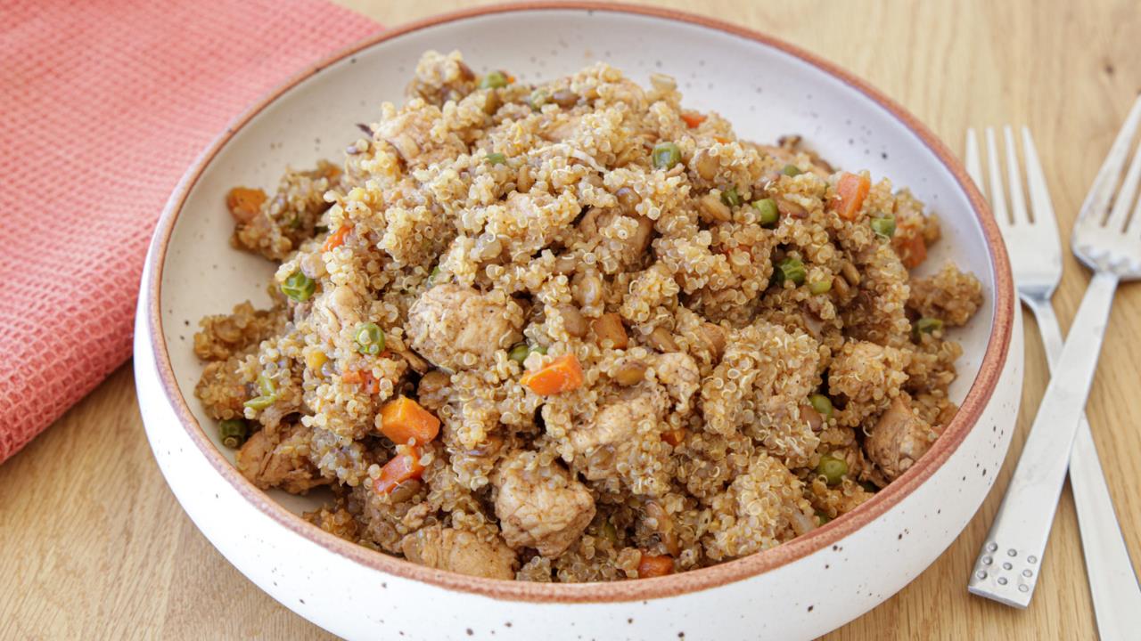 A bowl of quinoa fried rice with mixed vegetables like carrots, peas, and diced chicken. The dish is served in a speckled ceramic bowl placed on a wooden table, with a pink cloth napkin and silver utensils beside it.