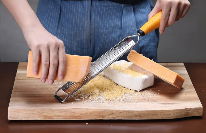 Hands are grating a block of cheese on a fine grater over a wooden cutting board. The person is wearing a blue apron. There is a smaller block of cheese and a white block of cheese also on the cutting board. Grated cheese is scattered around.