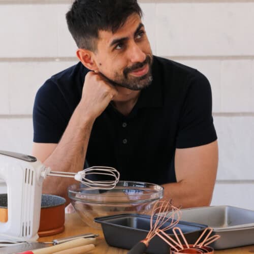 A man with short dark hair and a beard is sitting at a wooden table, resting his head on one hand, and looking to the side. The table is covered with various kitchen utensils, including a mixing bowl, whisks, a hand mixer, and baking pans.