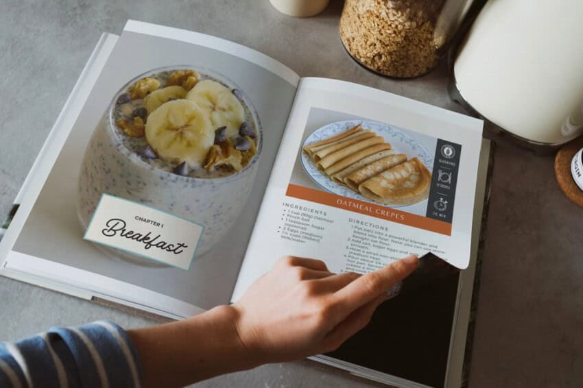 A person is flipping through a cookbook open to a page with breakfast recipes. The left page features a bowl of oatmeal topped with bananas and nuts. The right page shows a recipe for oatmeal crepes, with a photo of the crepes and a list of ingredients and directions.