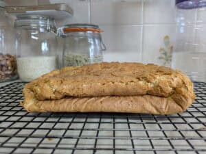 A loaf of bread with a sunken, collapsed top sits on a cooling rack in a kitchen, with jars of dry ingredients visible in the background.