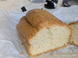 A loaf of homemade bread, partially sliced, sits on parchment paper. The bread has a golden-brown crust and a dense, light-colored interior. The background shows a blurred countertop and kitchen items.